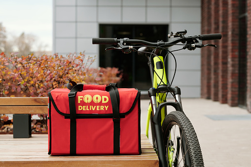 Bicycle of courier of food delivery service standing by wooden bench with big red square backpack containing online orders of clients