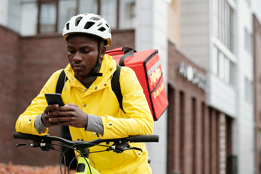 Serious black man in safety helmet and uniform of worker of food delivery service sitting on bicycle and checking new online orders on phone