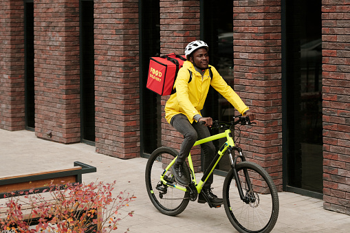 Deliveryman of modern food delivery service with orders of online clients in big red backpack moving along brick building on bicycle