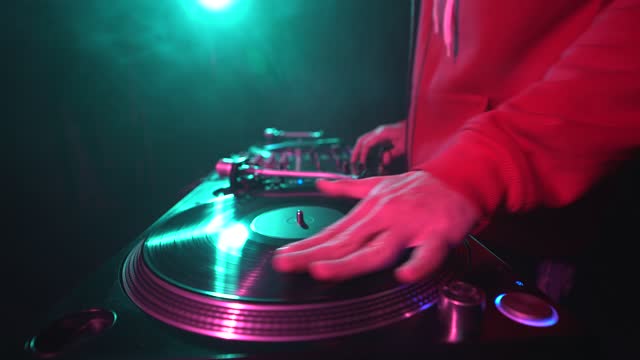 Hip hop DJ in red hoodie scratches vinyl record disc on turntable player in close up wide angle video. Club disc jockey scratching records on party