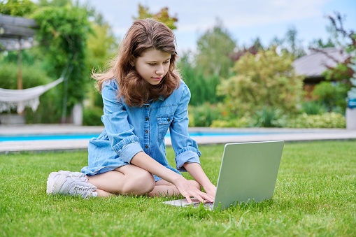 Preteen girl sitting on backyard lawn using laptop for leisure and study. Childhood, adolescence, lifestyle, education concept