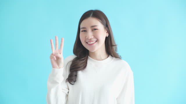 Young asian woman smiling and counting on her fingers up to 3, Happy asia woman looking at camera and showing one, two, three hand sign while standing over isolated blue background in studio
