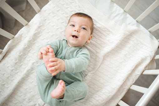 Funny smiling infant baby boy wake up in bed crib look at camera over white blanket top view closeup. Childhood. Good morning.