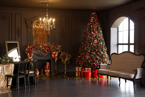 Interior of luxury dark living room with fireplace, comfortable sofa and chandelier decorated with Christmas tree and gifts.