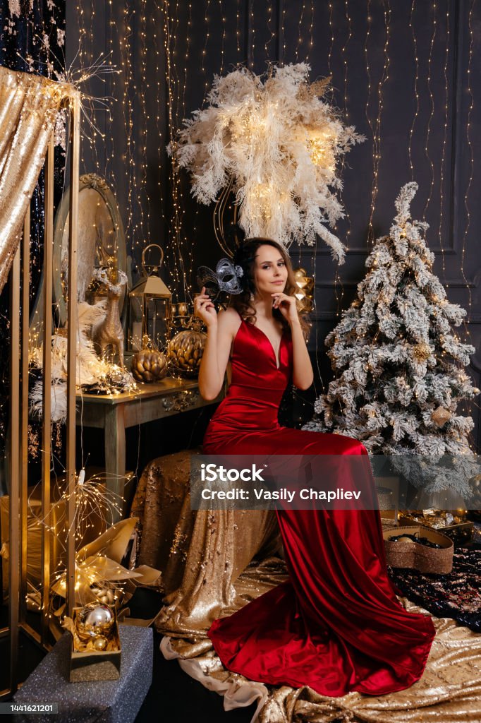 Foto de Uma Menina Com Cabelo Escuro Em Um Vestido Apertado De Seda Vermelha  Sentase Perto De Uma Penteadeira Uma Árvore De Natal E Decorações De Natal  Contra Uma Parede Preta e