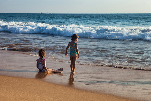 Rear view two happy little girls in swimsuits resting on sandy beach on tropical sea at waves background. Children from behind in summertime holiday. Travel vacation concept. Copy text space
