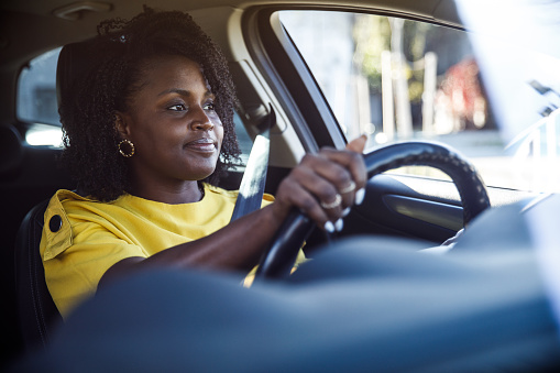 Portrait of charming young Black woman sitting in driver's seat in her car and smiling and enjoying the ride to her location. Seen through windshield.