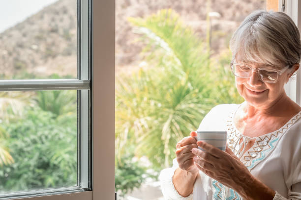 istock Smiling senior woman alone indoors standing at the window while holding a coffee cup. Elderly caucasian female enjoying a hot drink 1441630371