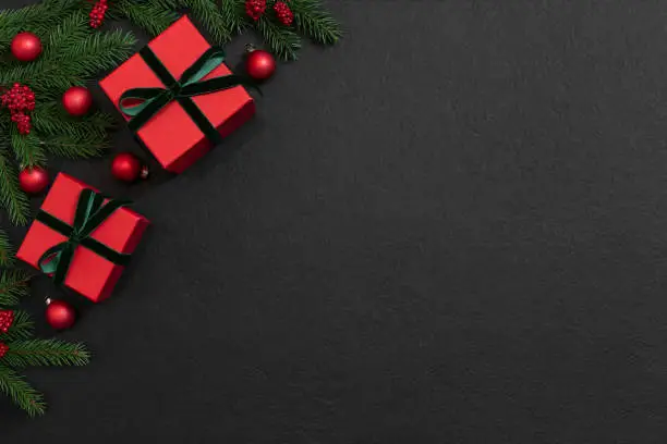 Photo of Christmas angle frame with red gift boxes, fir branches and festive decor on a black background. New Year border. Copy space, top view, flat lay.