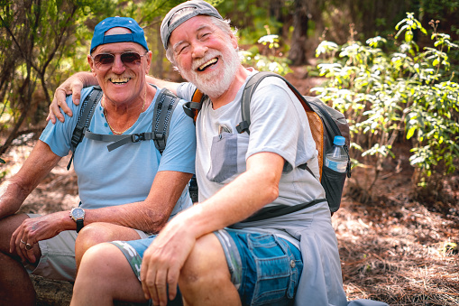 Couple of happy senior men in mountain excursion embracing looking at camera laughing while sitting to rest, enjoying healthy lifestyle in nature