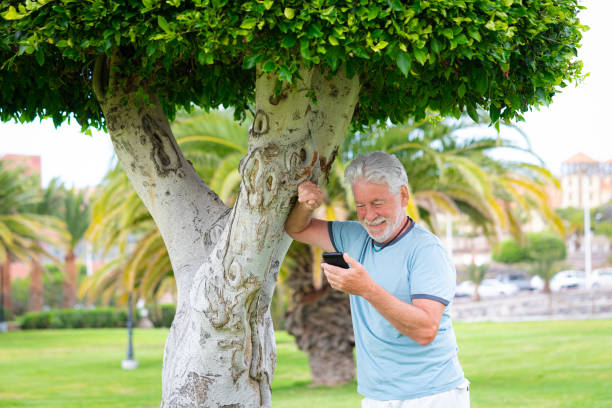 Beautiful bearded senior man standing under a tree in public park using mobile phone. Smiling elderly man holding telephone using app and social media