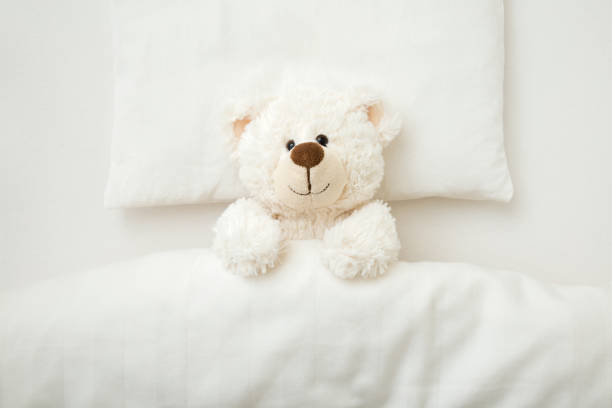 istock Adorable smiling white teddy bear lying down on pillow and sheet under blanket in baby crib at home. Closeup. Top down view. 1441638314