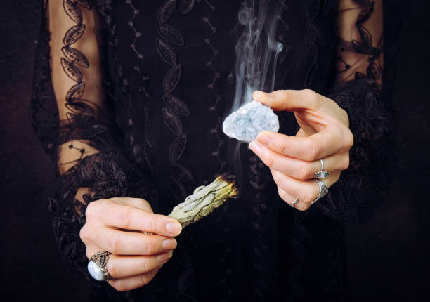 Close up view of woman in black lace dress, cleansing blue celestite  crystal cluster gemstone by smudging white sage bundle. Remove negative energy, cleanse crystal. stock photo