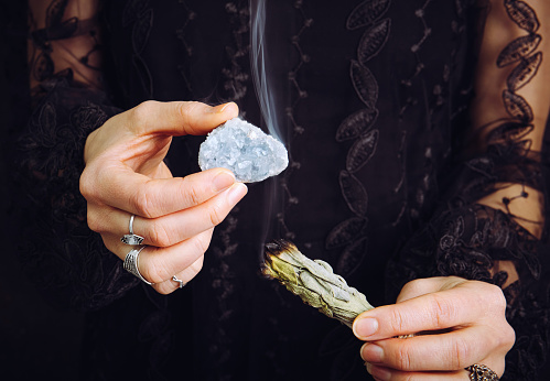Close up view of woman in black lace dress, cleansing blue celestite  crystal cluster gemstone by smudging white sage bundle. Remove negative energy, cleanse crystal.