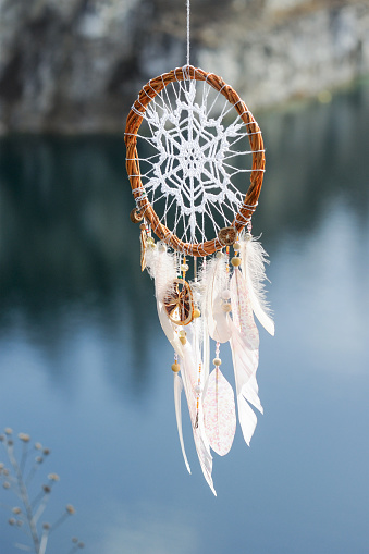 Handmade pink dream catcher with white doily on background of rocks and lake. Tribal elements, feathers, lace, crochet snowflake