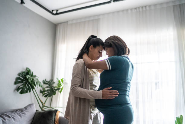 Lesbian couple embracing in the living room at home - including a pregnant mid adult woman