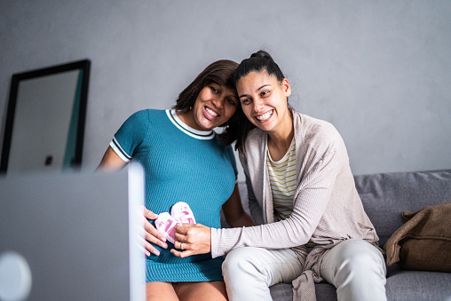Lesbian couple showing a baby shoe in a video call in the living room at home - including a pregnant mid adult woman
