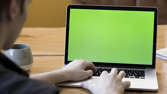 Rear view of a man typing on the laptop with green screen, sitting at the wooden table with a big cup of tea or coffee. Male working at his desk at the computer, chromakey.
