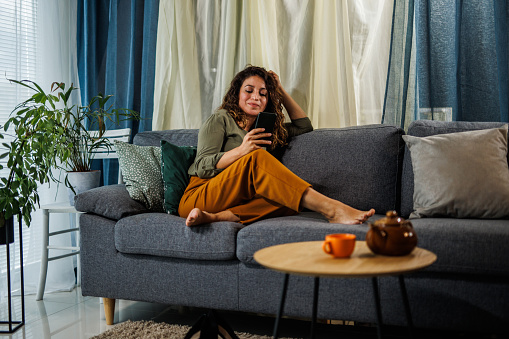 Wide shot of contented mid adult woman relaxing on the cozy sofa in her living room, scrolling through social media via smart phone and resting after coming home from work.