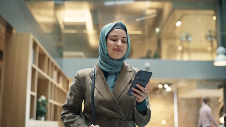 Portrait of Female Muslim Lecturer Leaving Campus While Browsing on her Smartphone. Hijabi Young Woman Smiling After Successfully Finishing a Class and Receiving Positive Feedback from Students