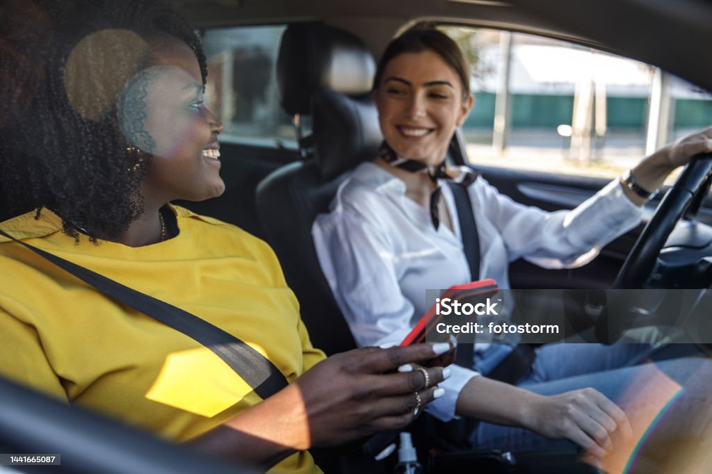 Young woman driving while her frined is looking up direction on a map via smart phonepassenger seat Selective focus shot of smiling young woman sitting in driver's seat in her car and driving while her friend in a passenger seat is looking up directions via smart phone. Car Pooling Stock Photo