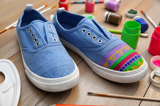 Light blue sneakers and painting supplies on wooden table, closeup. Customized shoes