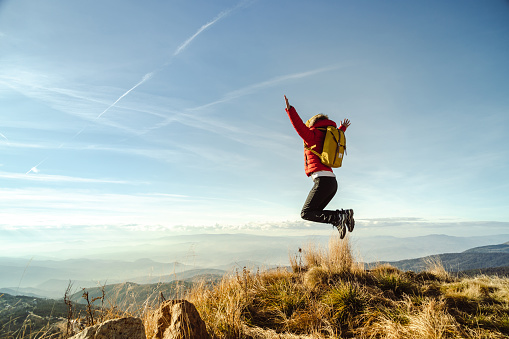 Young female hiker with a backpack jumping on top of a mountain and looking at the beautiful view