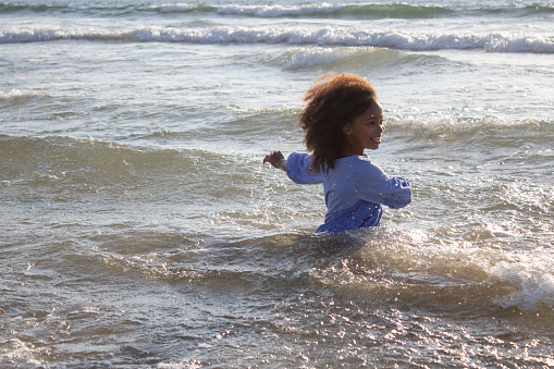 Brave little girl in dress on beach. African American child on beach on summer day, splashing water, smiling, playing in waves. Childhood, vacation, happiness concept