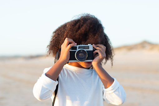 Portrait of cute girl with old-fashioned camera. Female model with curly hair and in white pullover holding camera on beach. Childhood, hobby concept