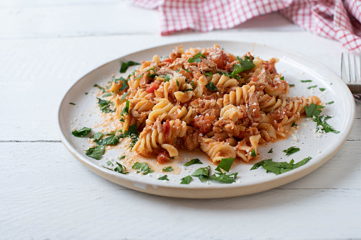 Delicious homemade, italian pasta with tuna, tomato sauce and parmesan cheese. Served on a plate isolated on white wooden table background with copy space. Front view