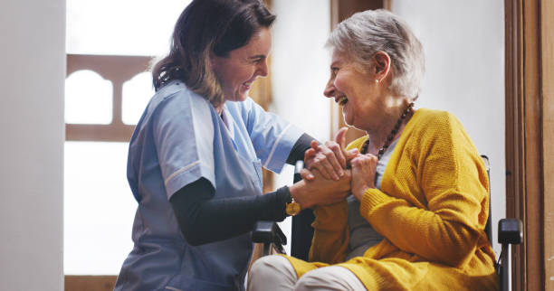 Nurse, laughing and senior woman in nursing home bonding, smiling and holding hands. Healthcare worker laugh with retired elderly woman in wheelchair sitting together for support, friendship and care stock photo