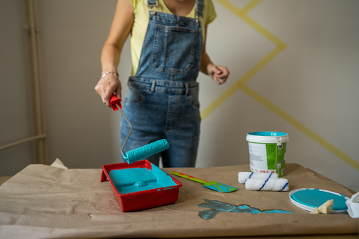 Unrecognizable woman decorating her living room, she is dipping paint roller in paint before painting a wall.