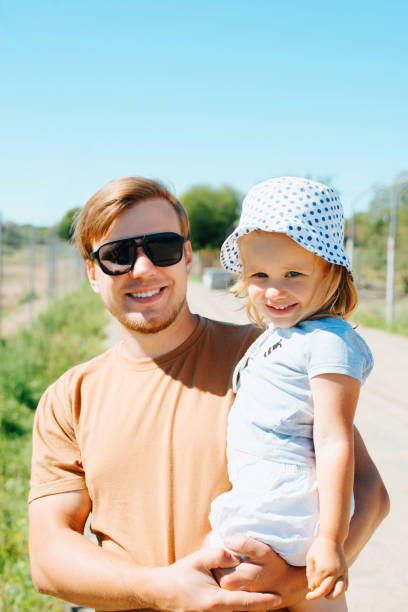 Portrait of dad with daughter outdoors. Young smiling father in sunglasses holding a baby in arms and looking at camera on a sunny day stock photo