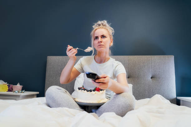 istock Emotionally stressed, shocked woman eating cake and holding a remote control while watching TV in bed. Scary horror movie or terrible news, scare in her eyes Overeating, Jamming of negative emotions. 1441685343