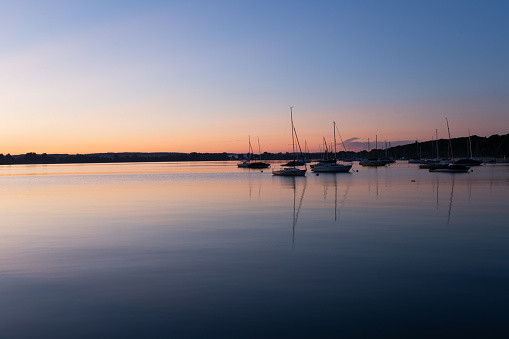 Several sailing ships are quietly anchored on the shores of the Ammersee. The golden hour is just turning into the blue hour and twilight is advancing. Panorama shot in wide angle.