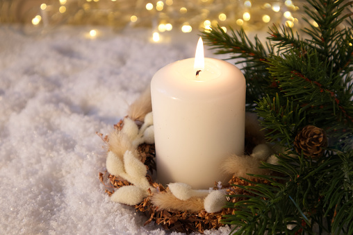 A white candle decorated with Christmas decor on the snow against the background of lights
