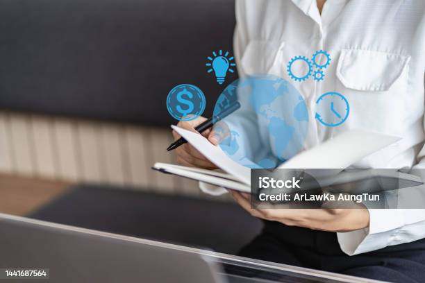 Young Asian College Student Reading A Book Doing Homework And Using Technology Laptop In University Library With Money Icons And Light Bulb Clock Stock Photo - Download Image Now