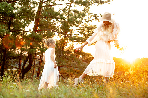 Mother run and lead by hand little daughter through coniferous wood at yellow sunset, sun shining. Mom and child play in forest at summer together in evening, family white look, picturesque landscape.