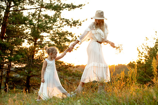 Mother lead by hand little daughter through coniferous wood at sunset, sky background. Mom and child walk in forest at summer together on green grass, family white look, picturesque landscape.