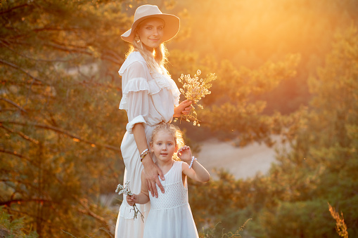 Mother with cute little daughter stand on high hill in white dresses in wood at yellow sunset. Mom and child walk in forest at summer evening with bouquet of wildflowers, family look, landscape.
