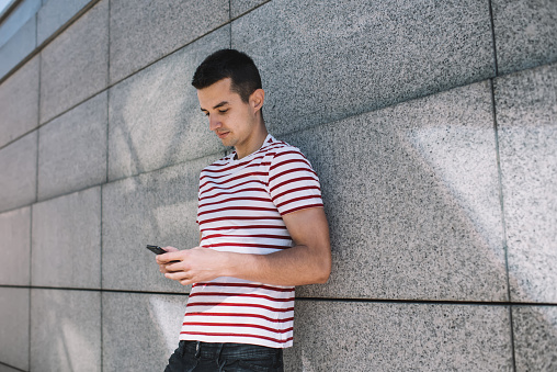 Low angle of young man surfing Internet on mobile phone and leaning on concrete building while strolling in city at daytime