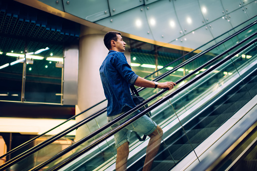 Low angle side view of young male in casual outfit going up on escalator at modern building with glass walls