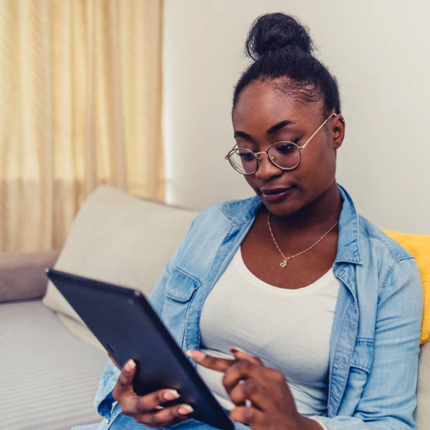 istock Happy young Black girl relaxing on sofa, using tablet computer, reading book, watching movie, media content, chatting online, talking on video call, laughing, smiling. Internet communication. 1441693272