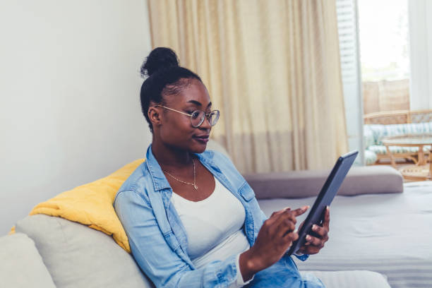 istock Smiling woman working online with a digital tablet while relaxing on a sofa in her bright living room at home. 1441693523