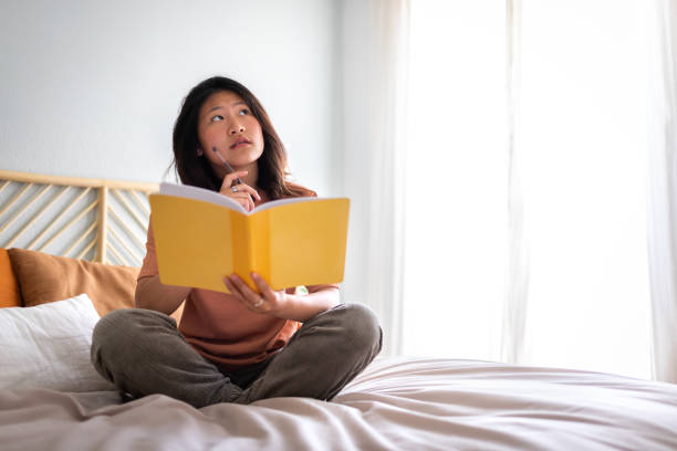 istock Teen asian girl sitting on bed thinking, using diary and pen to write down thoughts and ideas. Copy space. 1441701419