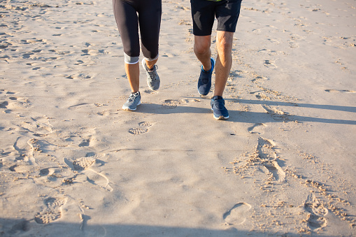 Unrecognizable man and woman in sneakers running on sandy beach. Close-up shot of mature couple in sportswear having cardio training on summer day, taking care of health. Outdoor activity concept