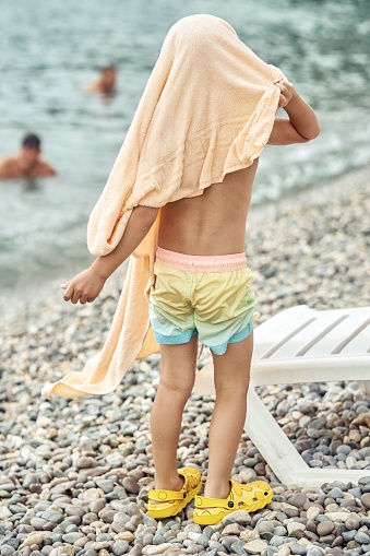 Preschooler boy wearing beige terry towel on head stands on pebble beach exploring sea. Child enjoys walking on beach near beach chair by sea water