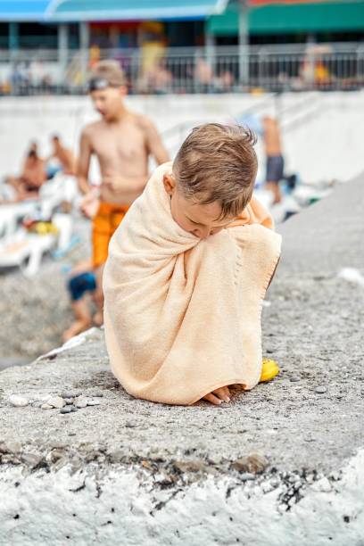 preschooler boy in towel sit on pier gets cold after swimming in sea - beach stone wall one person imagens e fotografias de stock