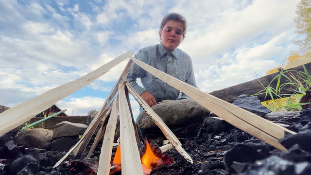 An Elementary-Aged Caucasian Boy Adds Larger Pieces of Kindling Wood to a Fire Teepee while Building an Outdoor Campfire