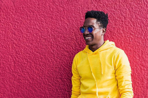 Portrait of a smiling young African man wearing a yellow sweatshirt and sunglasses in front of a pink or purple rough wall.